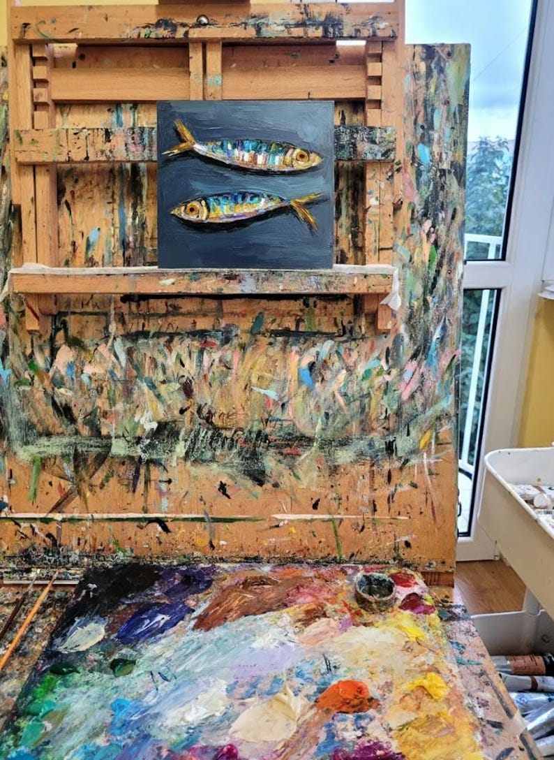 Oil painting of two fish with colorful scales and yellow fins on a dark blue background. The artwork is displayed on an artist's easel, surrounded by paint splatters and a colorful palette, suggesting a studio setting.