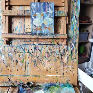 May include: An original oil painting on canvas of two white ducks with orange feet, set against a blue and white background. The artwork is displayed on a wooden easel covered in paint splatters, suggesting an artist's studio.