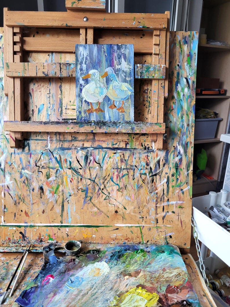May include: An original oil painting on canvas of two white ducks with orange feet, set against a blue and white background. The artwork is displayed on a wooden easel covered in paint splatters, suggesting an artist's studio.