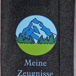 May include: A dark gray felt document holder with a circular graphic of a mountain scene in blue, green, and white. The text "Meine Zeugnisse" is printed in light blue below the graphic.