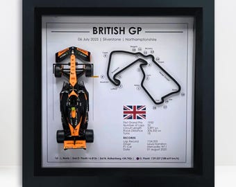 Può includere: Opera d'arte incorniciata con una vettura di Formula 1 nera e arancione, una mappa del circuito e dettagli del Gran Premio di Gran Bretagna. Il testo include data, luogo e statistiche della gara.