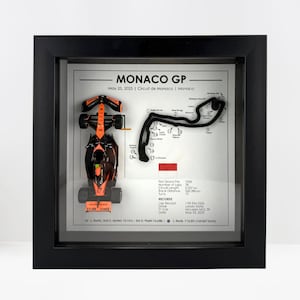 Puede incluir: Obra de arte enmarcada con un coche de carreras naranja y negro en miniatura y un diseño de circuito. La obra de arte incluye el texto "MONACO GP" y detalles de la carrera. El marco es negro.