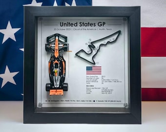 Puede incluir: Una vitrina enmarcada que presenta un coche de carreras de Fórmula 1 en miniatura con detalles en naranja y negro. El marco también incluye un esquema negro del circuito de las Américas con curvas numeradas. El texto "United States GP" y "22 de octubre de 2023" es visible en la parte superior del marco.