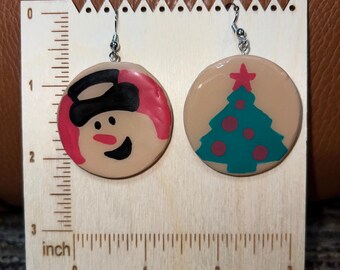 May include: A pair of Christmas-themed earrings. One earring features a snowman with a black hat and a pink scarf. The other earring has a teal Christmas tree with red star. Each earring is approximately 1 inch in diameter.