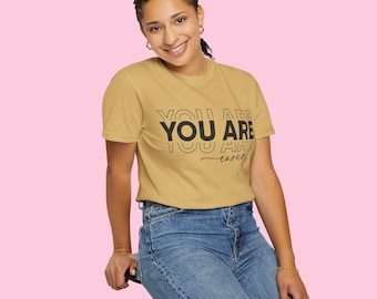 May include: Mustard yellow t-shirt with the text "YOU ARE enough" in black. The model is wearing blue jeans and is sitting on a black stool. The background is a solid pink color.