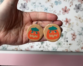 May include: A pair of round earrings featuring a pumpkin design. Each earring has a tan base with an orange pumpkin face and a green stem. The earrings are held up by silver hooks, perfect for Halloween.
