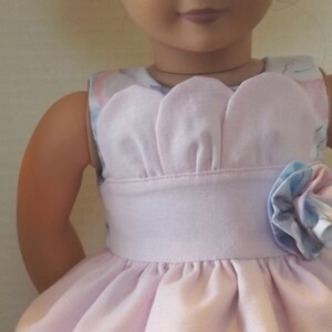 以下が含まれることがあります： フリルネックと花柄のディテールが施されたピンクのドレス。ドレスはノースリーブで、スカートはボリュームがあります。