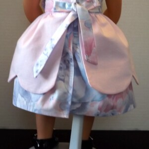 以下が含まれることがあります： 後ろに白いリボンが付いたピンクとブルーのフローラルドレス。ドレスは人形用で、フリルのある裾です。