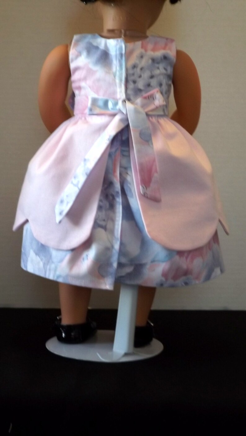以下が含まれることがあります： 後ろに白いリボンが付いたピンクとブルーのフローラルドレス。ドレスは人形用で、フリルのある裾です。