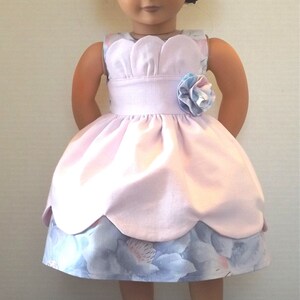 以下が含まれることがあります： 花柄のアクセントが付いたピンクとブルーのドレスを着た人形。人形は黒髪で緑色の目。人形は黒い靴を履いています。