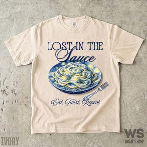 May include: An ivory short-sleeve t-shirt featuring a graphic of spaghetti on a blue plate with yellow swirls. The text reads LOST IN THE SAUCE above the plate and Eat, Twirl, Repeat below. The design mimics a famous post-impressionist painting style.
