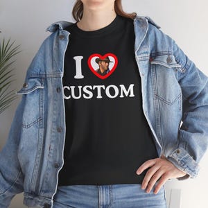 May include: Black t-shirt with the text "I ❤️ CUSTOM" in white. The heart symbol contains an image of a person wearing a hat. The person is wearing a denim jacket.