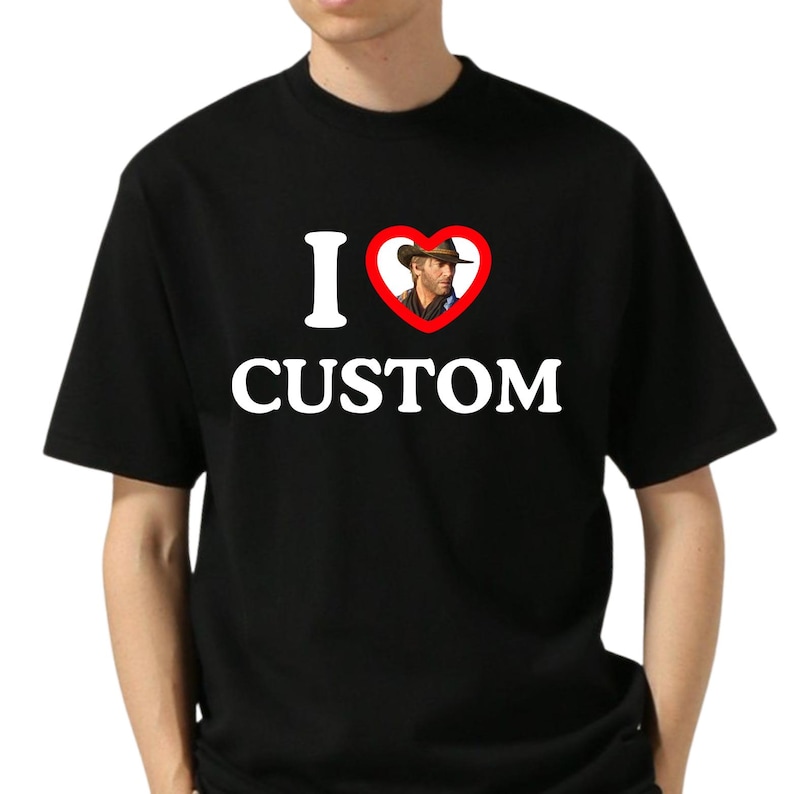 May include: Black t-shirt with the text "I &hearts; CUSTOM". The heart symbol contains an image of a man wearing a cowboy hat. The text and heart graphic are in white.