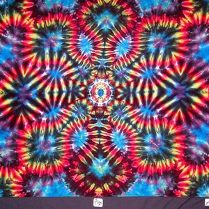 May include: A colorful tie-dye fabric with a symmetrical pattern of red, yellow, blue, and black swirls. The fabric has a central design with a smaller circle in the middle.