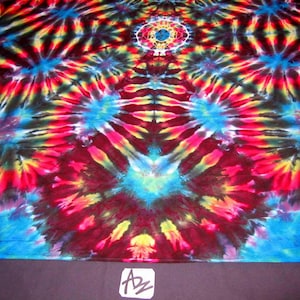 May include: A colorful tie-dye fabric with a circular pattern in shades of red, yellow, blue, and black. The fabric has a symmetrical design with a central circle and radiating lines.