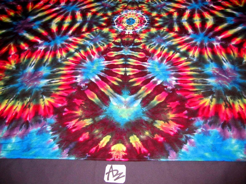 May include: A colorful tie-dye fabric with a circular pattern in shades of red, yellow, blue, and black. The fabric has a symmetrical design with a central circle and radiating lines.