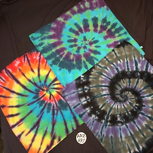 May include: Four square tie-dye bandanas in vibrant colors. One features a rainbow spiral, another a teal and purple spiral, a third a black and olive green spiral, and the last a teal and green spiral. The bandanas are arranged on a dark background.