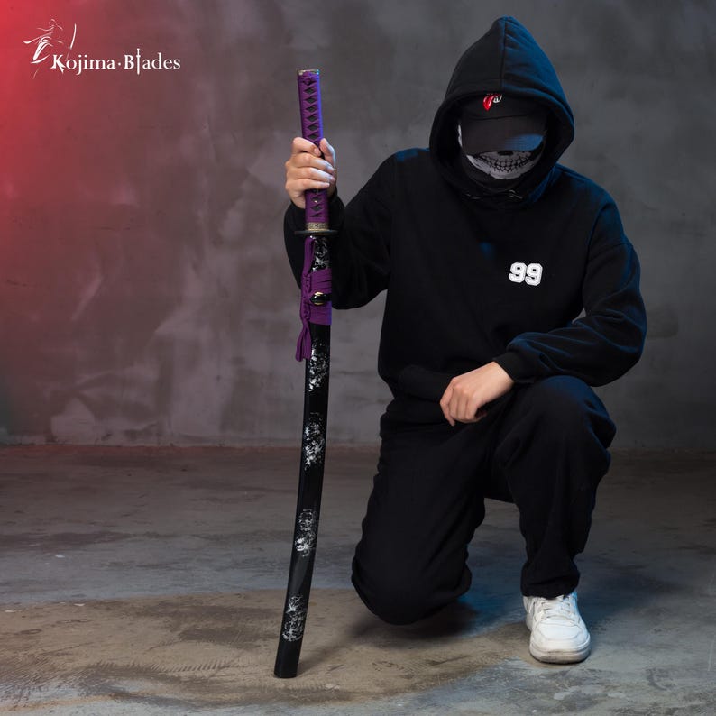 Pu&ograve; includere: Una persona indossa una felpa con cappuccio nera con il numero "99" sul davanti, una maschera facciale nera a forma di teschio e un berretto nero e bianco. La persona tiene in mano una katana nera con un'impugnatura viola e un design a forma di teschio bianco.
