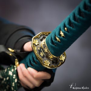 May include: Close-up of a teal and black sword handle with a gold-colored metal accent featuring a floral design. The handle is wrapped in a teal cord.