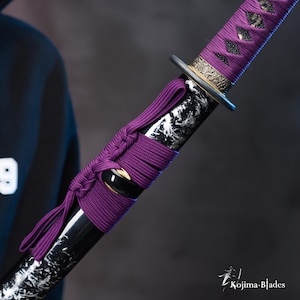 Pu&ograve; includere: Primo piano di una spada katana nera e bianca con un'impugnatura intrecciata viola. La spada ha un raccordo in metallo dorato nella parte superiore dell'impugnatura.