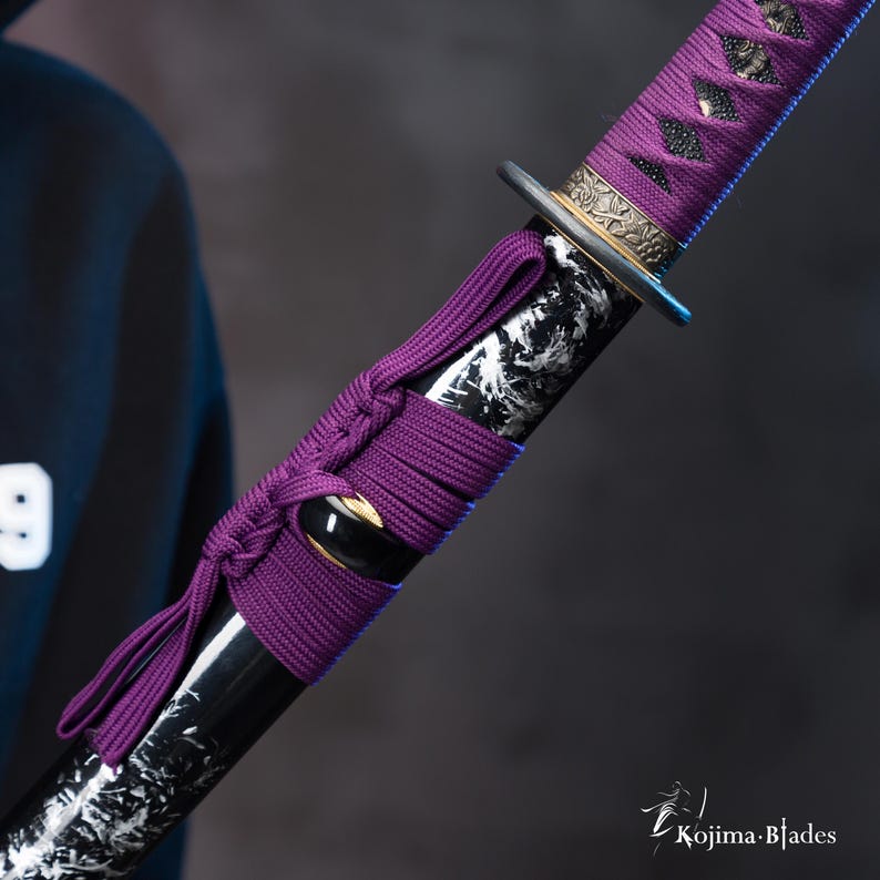 Pu&ograve; includere: Primo piano di una spada katana nera e bianca con un'impugnatura intrecciata viola. La spada ha un raccordo in metallo dorato nella parte superiore dell'impugnatura.