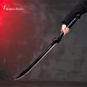 Könnte beinhalten: Ein schwarzes Schwert mit einem Wolfskopf als Griff. Das Schwert wird von einer Hand in einem schwarzen Ärmel gehalten. Der Hintergrund ist eine rote und graue Wand.