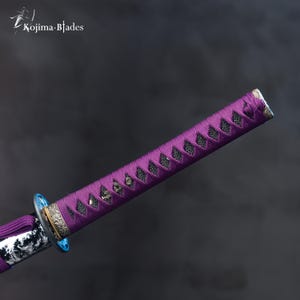 Pu&ograve; includere: Primo piano di un'impugnatura di katana viola con un rivestimento a motivi bianco e nero. L'impugnatura &egrave; decorata con un pezzo di metallo argentato e un accento blu.