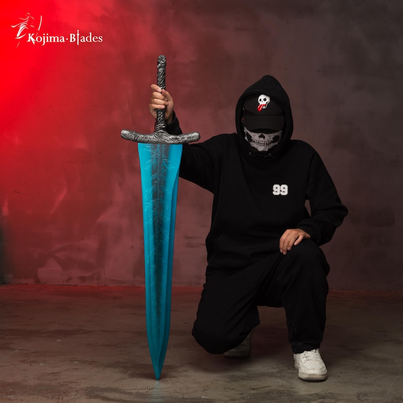 Op de afbeelding: Een persoon draagt een zwarte hoodie met een schedelgezichtsmasker en een zwarte pet met een witte schedel en een rode tong grafiek houdt een blauw en zilveren zwaard vast. De hoodie heeft het nummer 99 op de voorkant gedrukt.