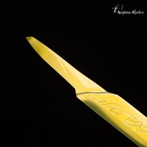 May include: A close-up of a yellow, sharp, pointed blade with the word "KOJIMA" etched into the side.
