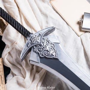 May include: A silver sword with a black, braided handle and intricate details on the pommel. The sword is lying on a white fabric surface.