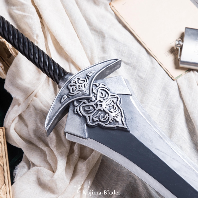 May include: A silver sword with a black, braided handle and intricate details on the pommel. The sword is lying on a white fabric surface.