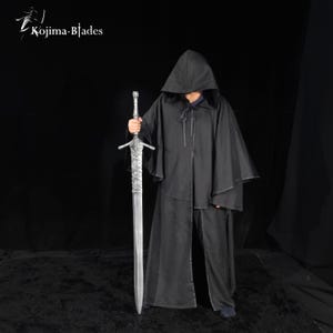 May include: A black hooded cloak with a front tie closure and a long silver-colored sword with a textured grip and crossguard. The Kojima Blades logo is visible in the top left corner. The sword features a metallic blade and a detailed hilt.