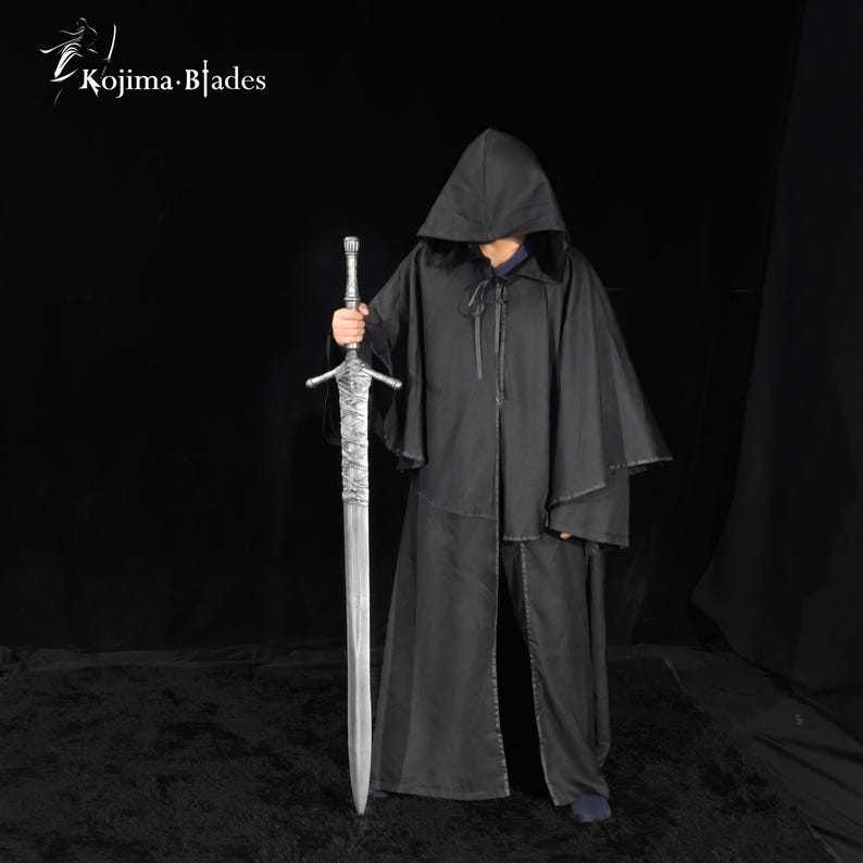 May include: A black hooded cloak with a front tie closure and a long silver-colored sword with a textured grip and crossguard. The Kojima Blades logo is visible in the top left corner. The sword features a metallic blade and a detailed hilt.
