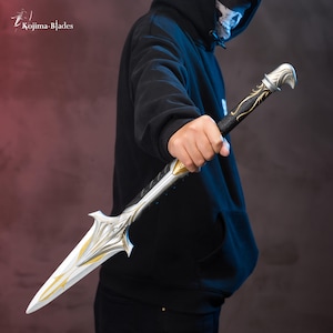 May include: A silver and gold-colored sword with a black handle. The sword has a sharp point and a decorative design on the blade. The sword is held by a person wearing a black hoodie.