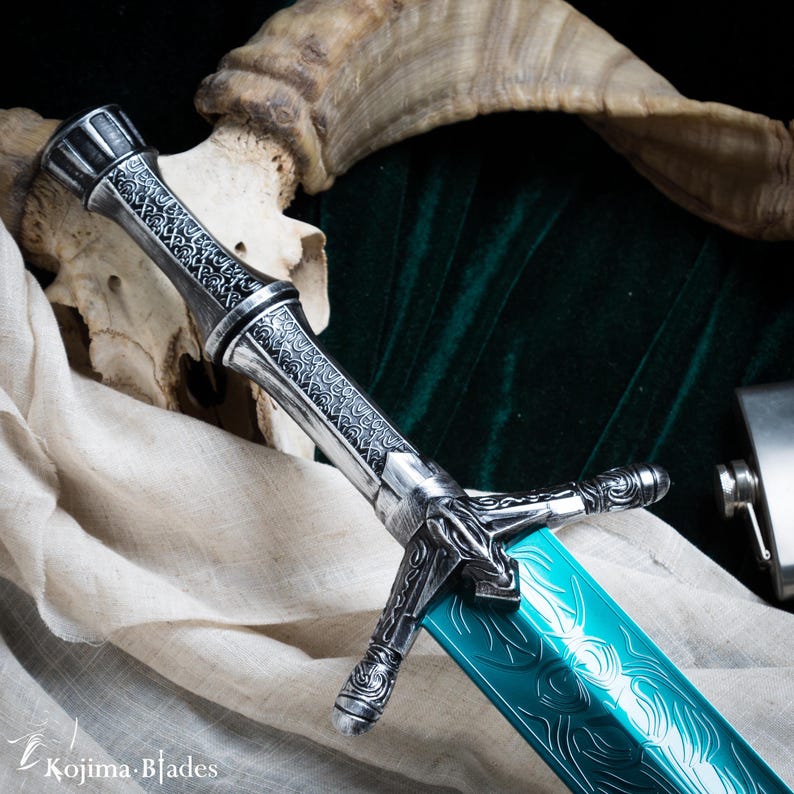 May include: A silver and teal sword with an intricate design on the handle and blade. The sword is resting on a white cloth and a skull.