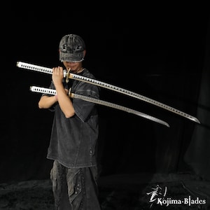Könnte beinhalten: Zwei Katana-Schwerter mit silberfarbenen Klingen und weiß umwickelten Griffen mit goldfarbenen Parierstangen. Die Schwerter werden horizontal vor der Brust gehalten. In der unteren rechten Ecke steht der Text Kojima Blades mit einem Schwert-Symbol.