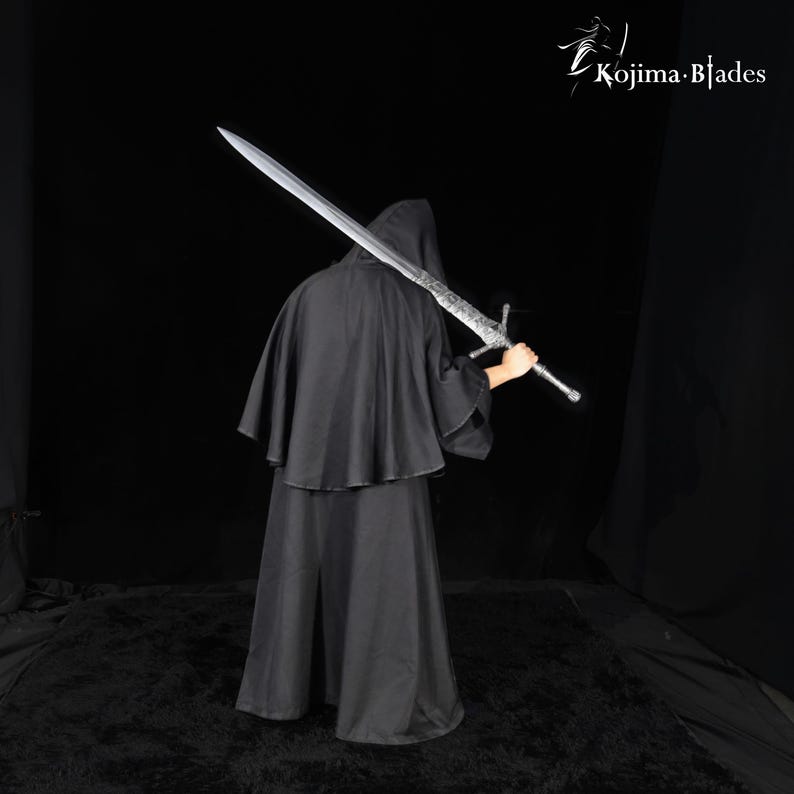 May include: A long, silver-colored metal sword with a detailed crossguard and pommel, held over the shoulder. The blade features etched markings near the hilt. The text Kojima Blades is visible in the upper right corner against a black background.