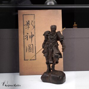 May include: A detailed wooden figurine of a warrior holding a spear, standing on a rock base. The figure is dark brown with intricate armor details. Behind it is a brown paper with Japanese characters. The Kojima-Blades logo is in the bottom left.