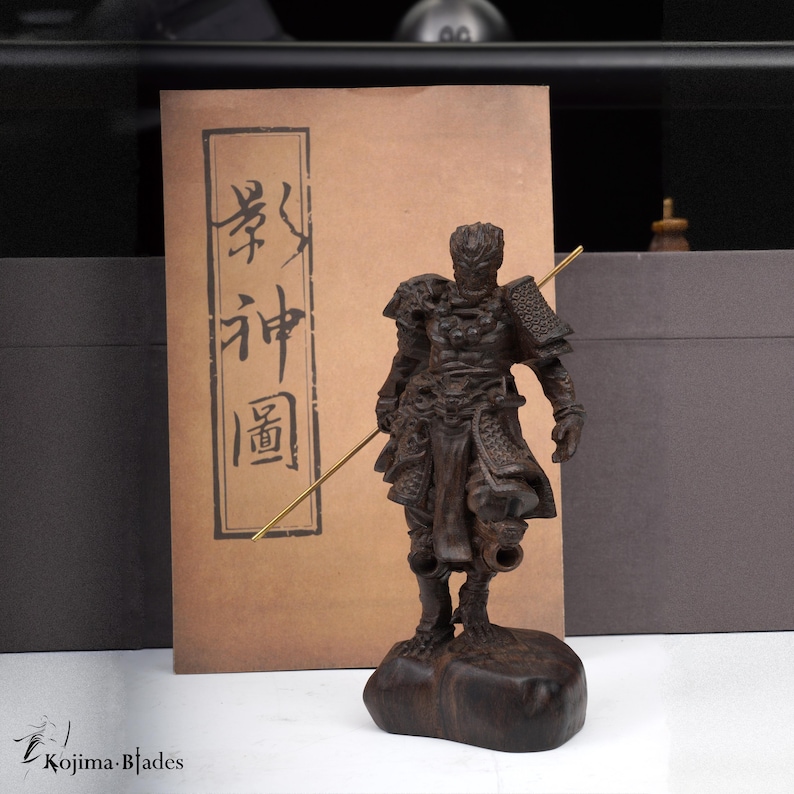 May include: A detailed wooden figurine of a warrior holding a spear, standing on a rock base. The figure is dark brown with intricate armor details. Behind it is a brown paper with Japanese characters. The Kojima-Blades logo is in the bottom left.