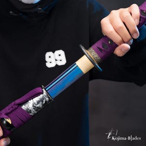 Puede incluir: Primer plano de una katana con mango morado y negro y hoja azul y morada. La espada la sostiene una persona que lleva una sudadera con capucha negra con el número 99 en la parte delantera.