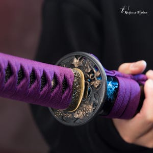 Pu&ograve; includere: Primo piano di un'impugnatura di spada viola e nera con lavorazioni in metallo intricate. L'impugnatura &egrave; avvolta da un cordoncino viola e presenta un pezzo di metallo circolare con un motivo floreale.