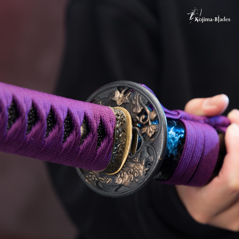 Pu&ograve; includere: Primo piano di un'impugnatura di spada viola e nera con lavorazioni in metallo intricate. L'impugnatura &egrave; avvolta da un cordoncino viola e presenta un pezzo di metallo circolare con un motivo floreale.