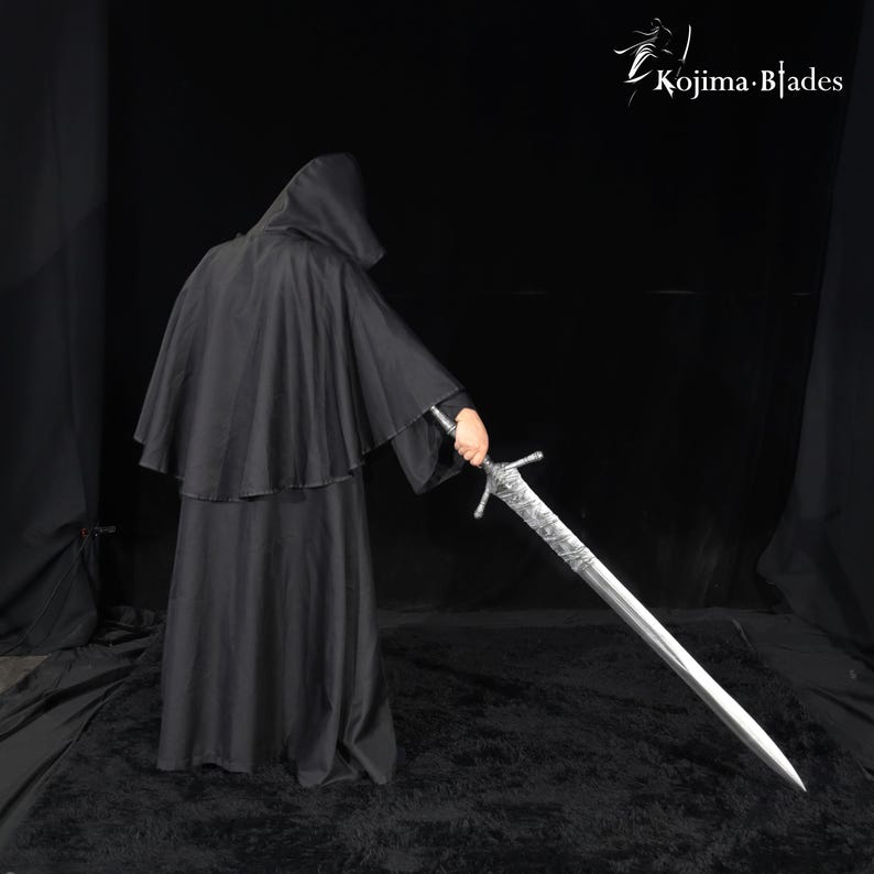 May include: A long, silver-colored metal sword with an intricate, textured hilt and crossguard, held by a figure wearing a black hooded cloak. The text Kojima Blades is visible in the upper right corner against a solid black background.