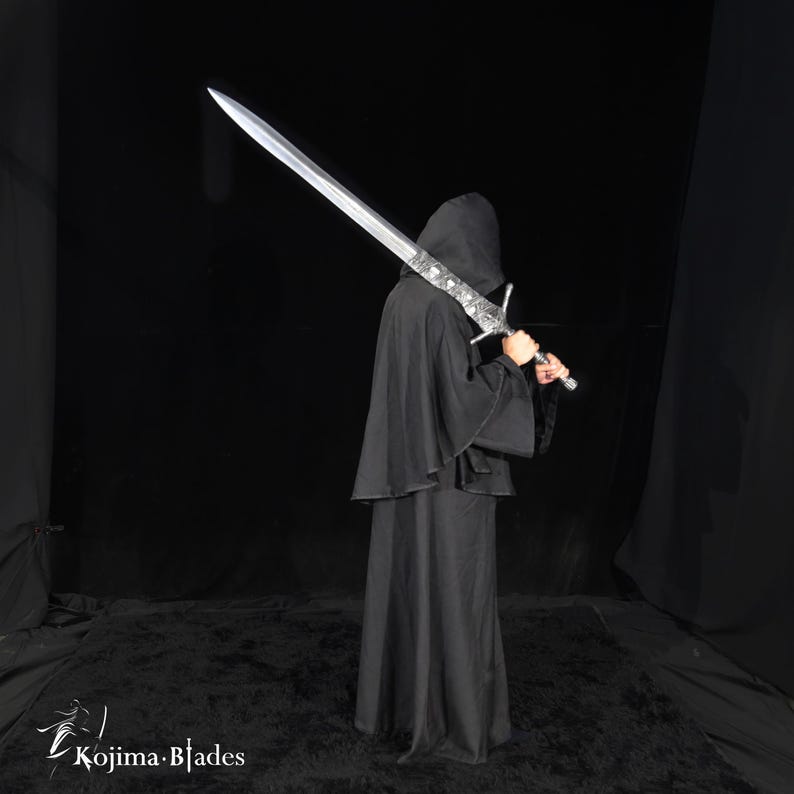 May include: A long, silver-colored metal sword with an ornate, detailed crossguard and hilt, held over the shoulder by a figure wearing a solid black hooded cloak. The blade is straight and pointed. The text Kojima Blades is visible.