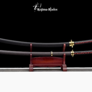 May include: Two black katana swords with red and black wrapped handles are displayed on a wooden stand. The swords are on a black background. The text "Kojima Blades" is visible in the top center of the image.