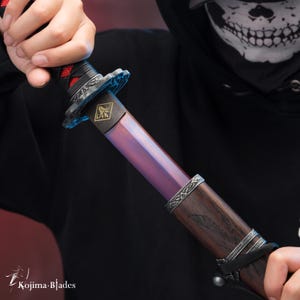 May include: A close-up of a katana sword with a dark wood handle and a purple and blue blade. The blade has a decorative design near the handle and a gold emblem on the blade.