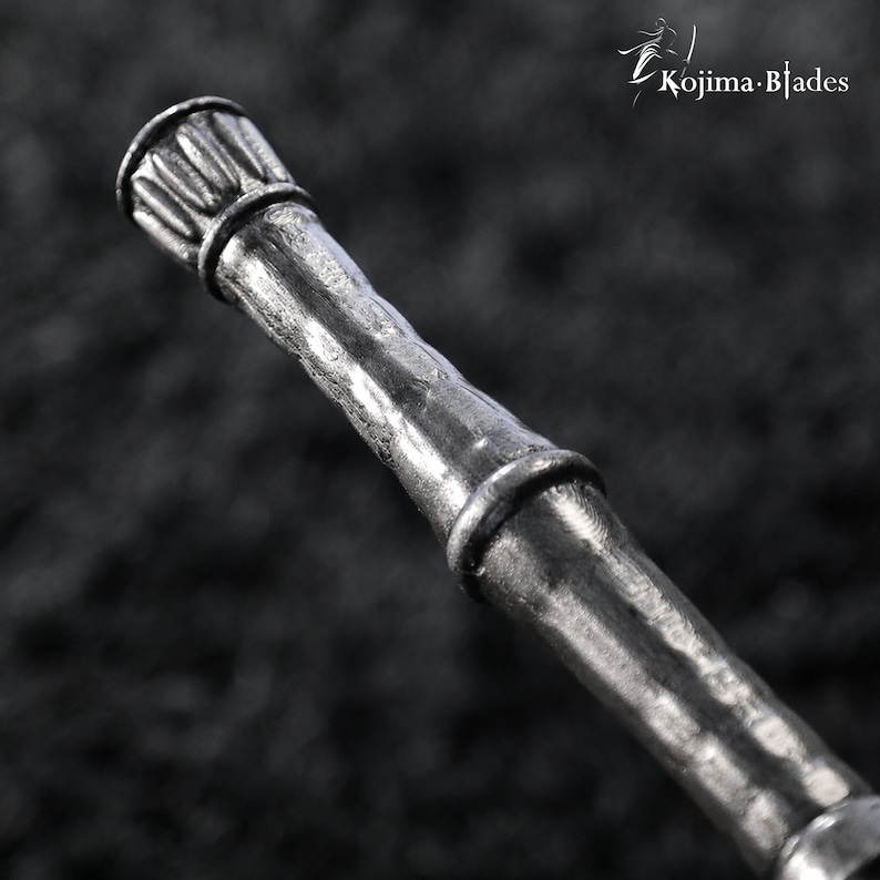 May include: A metallic silver-toned handle with a textured, bamboo-like appearance featuring segmented nodes and a fluted cap at the top end. The text Kojima Blades is visible in the upper right corner of the frame against a dark background.