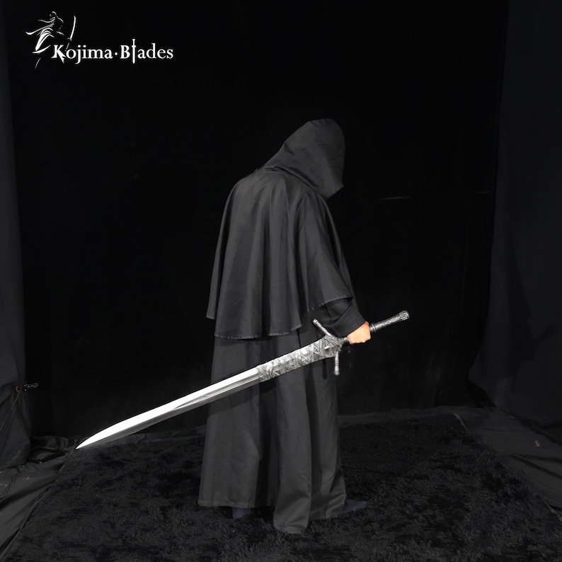 May include: A long, double-edged steel sword with an intricate, etched pattern on the blade near the hilt. The sword features a straight crossguard and a textured grip. The Kojima Blades logo is visible in the upper left corner of the image.