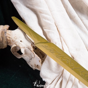 May include: A gold-coloured sword blade with intricate engravings. The blade is resting on a white cloth and a skull.