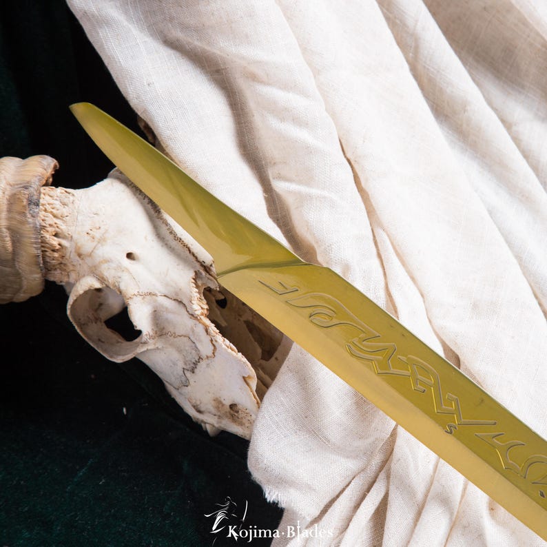 May include: A gold-coloured sword blade with intricate engravings. The blade is resting on a white cloth and a skull.